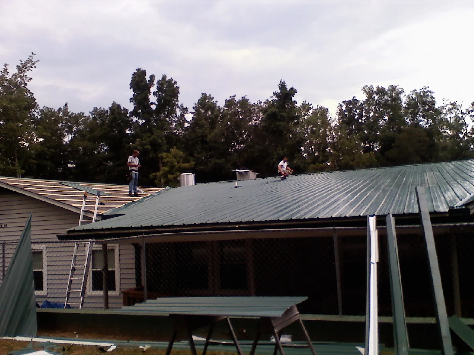 Two roofers installing green metal panels above a covered porch in Belmont